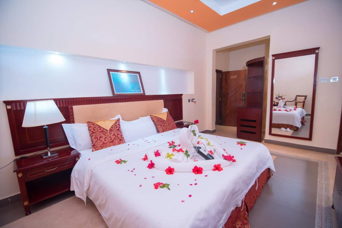 A hotel room with a large bed, a nightstand with a lamp, a wooden headboard, a framed picture on the wall, and a mirror on the door.
