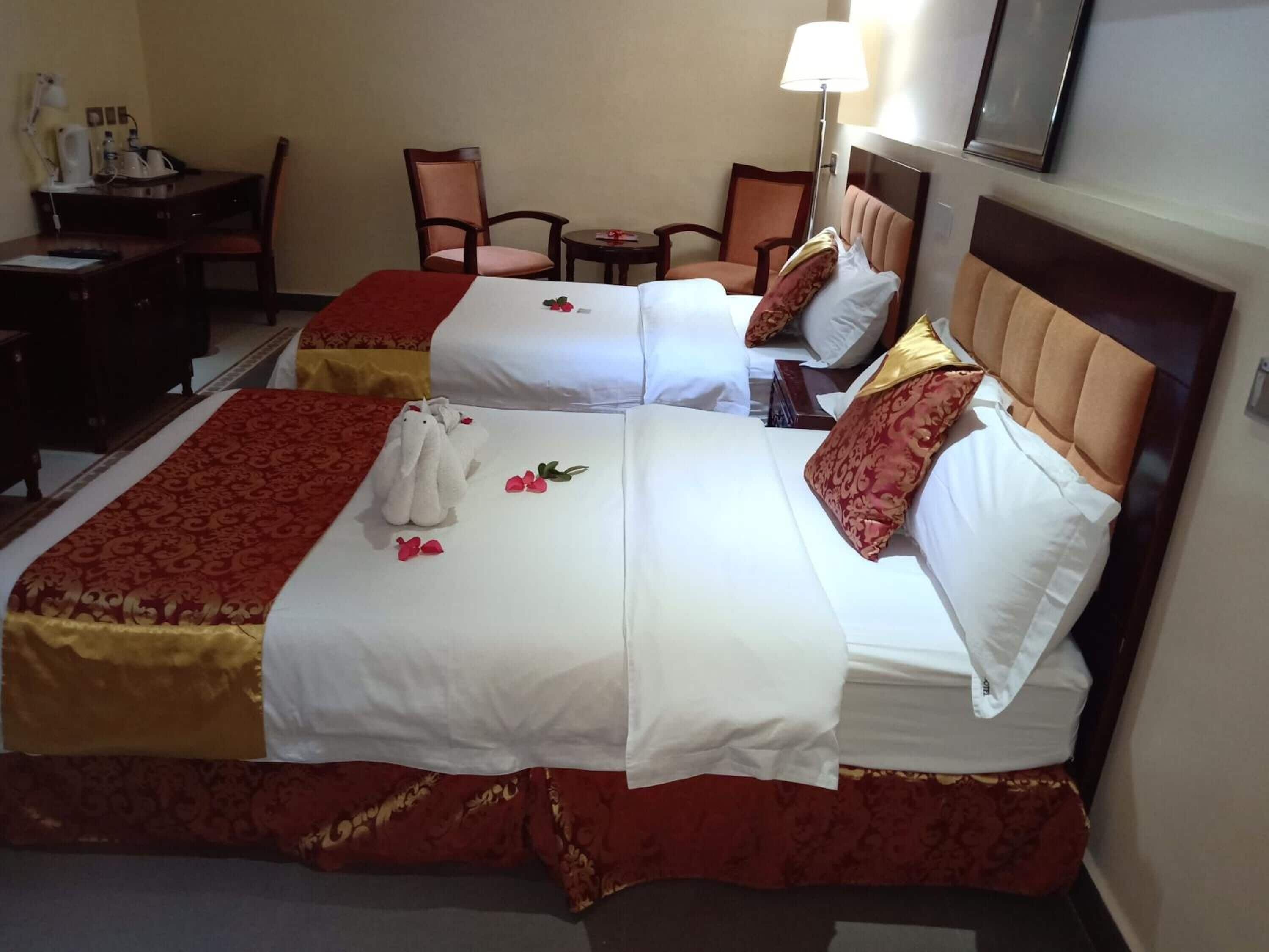 Une chambre d’hôtel avec deux lits, chacun recouvert d’une couvre-lit rouge et or, agrémentée de draps blancs et de coussins décoratifs. On y trouve un bureau avec une lampe, une chaise et une petite table également dotée d’une lampe.
