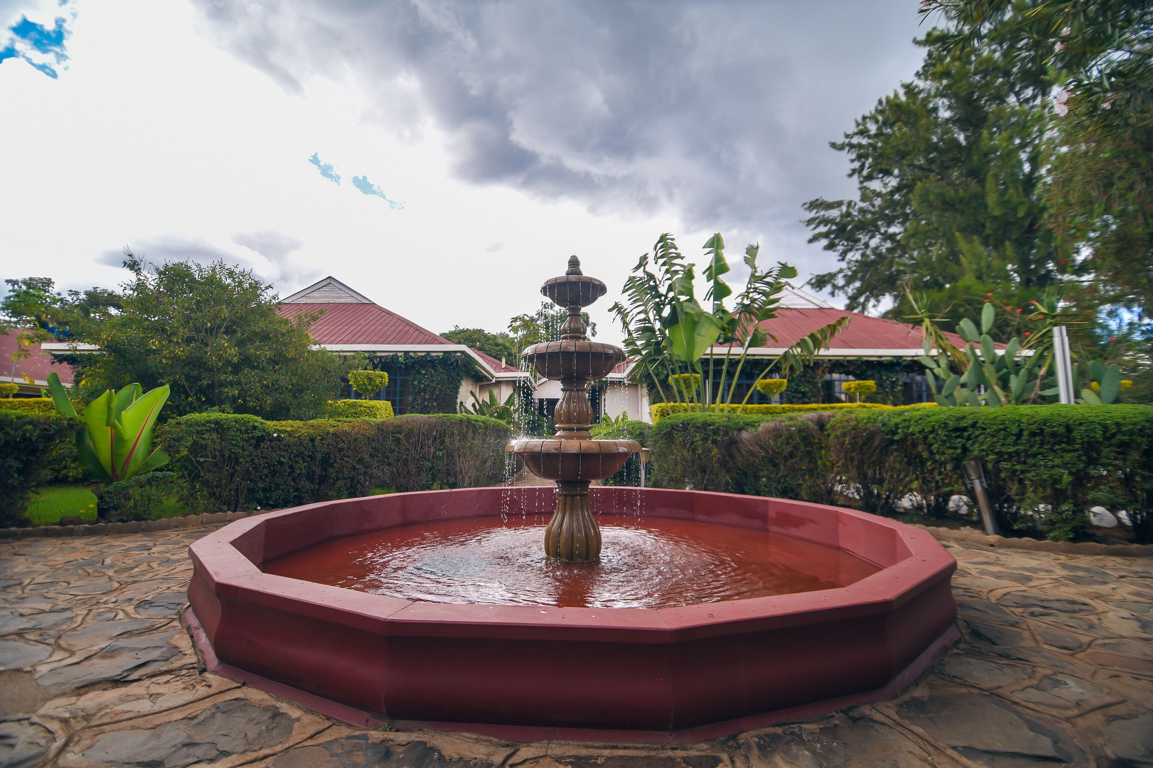 A stone fountain with water cascading in a circular basin, surrounded by neatly trimmed hedges and trees.