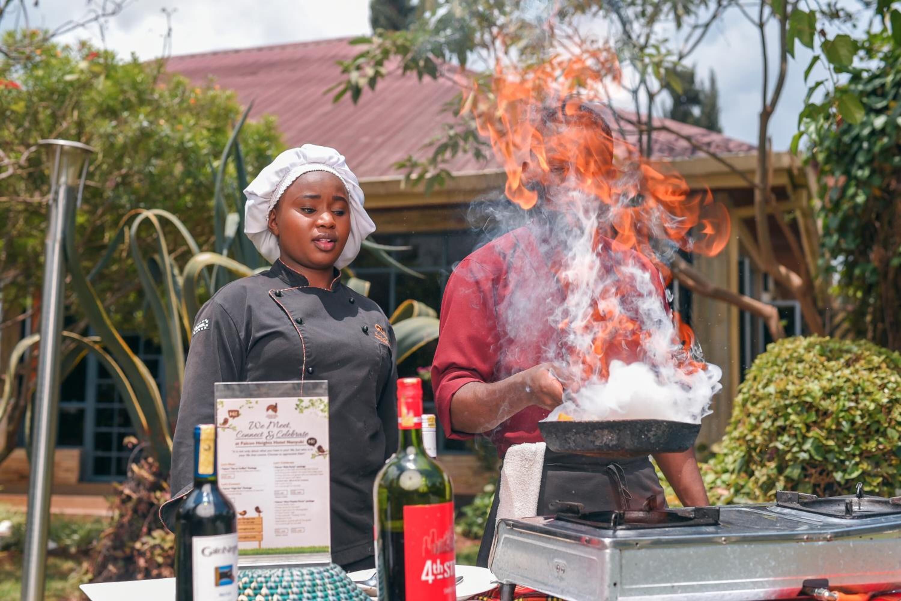A chef cooking outdoors with a grill, a menu stand, and a bottle of wine visible.
