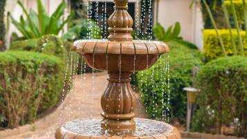 Una fuente escalonada con agua cayendo en cascada, rodeada de abundante vegetación y un edificio con techo rojo al fondo.