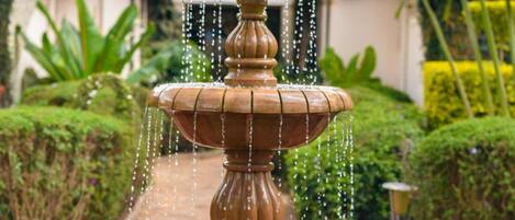 A tiered fountain with water cascading down, surrounded by lush greenery and a building with a red roof in the background.