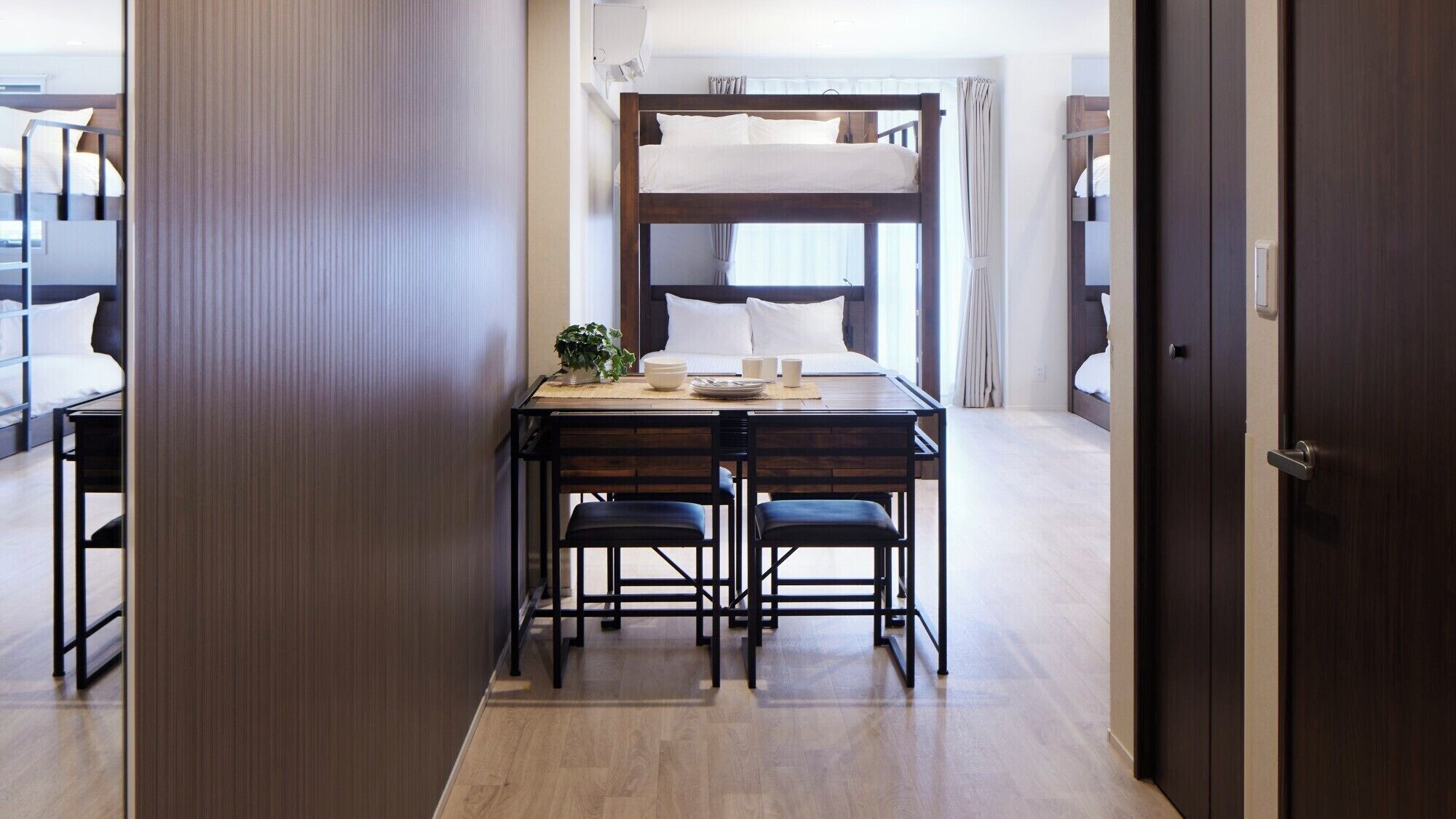 A hotel room with bunk beds, a wooden dining table with chairs, and a small table with a plant and cups.