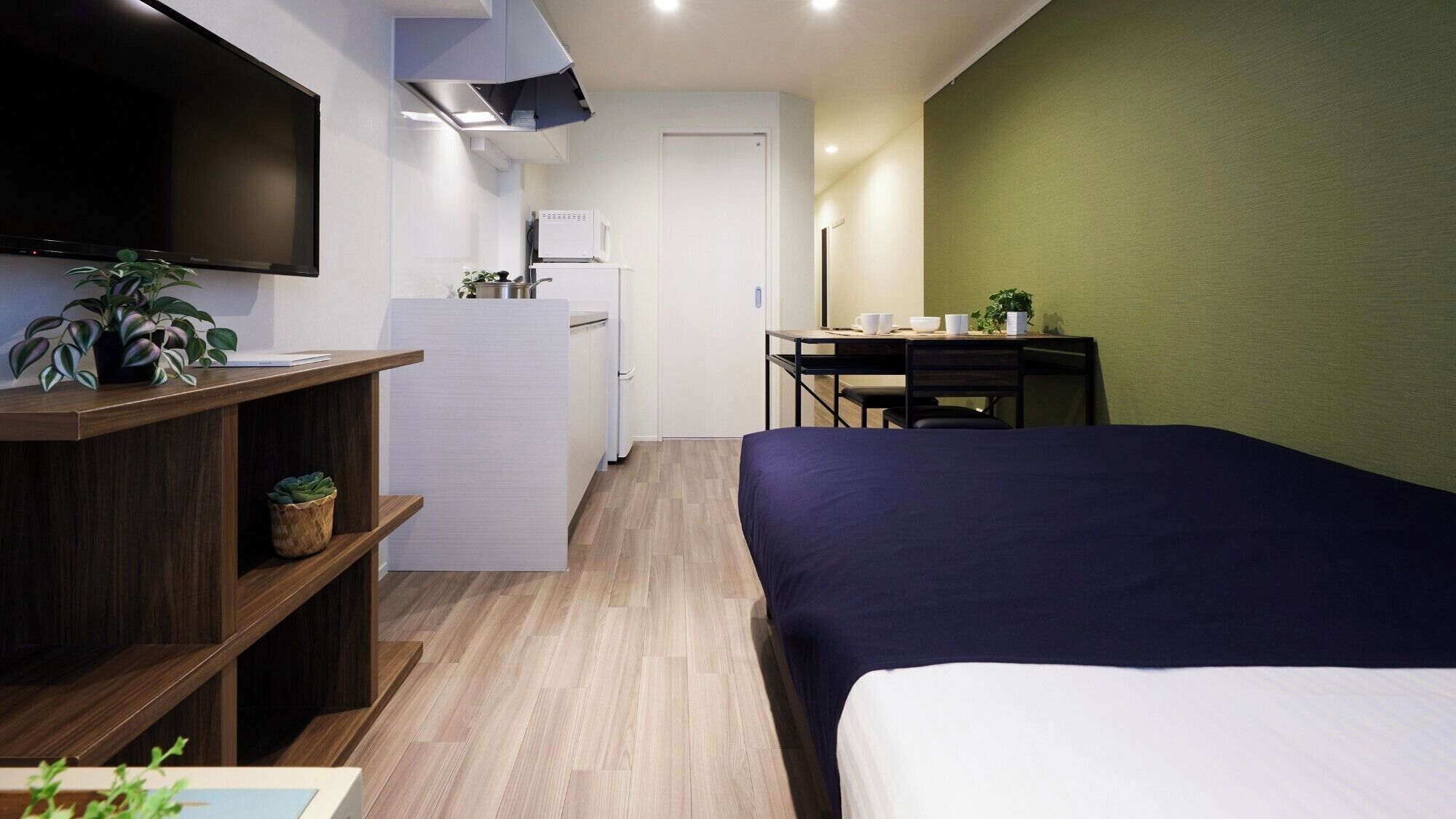 A compact hotel room with a bed, a desk, a television, a refrigerator, and a green wall.