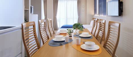 A dining area with a wooden table set for four, surrounded by wooden chairs. There is a bed with a blue bedspread in the background, a television mounted on the wall, and a window with curtains.
