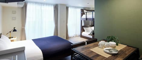 A hotel room with a bed, a desk with a chair, a small table with a plant, and a window with curtains.