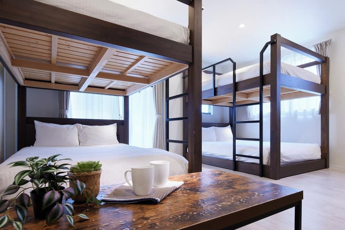 Habitación con literas, techo de madera, una cama individual con ropa de cama blanca, una mesita con una taza y una planta, y una ventana con cortinas.