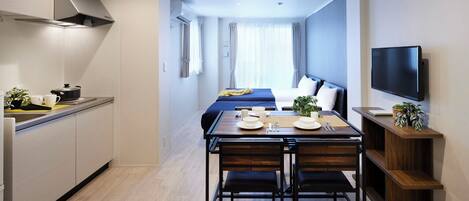 A compact hotel room with a kitchenette, a dining area with a table set for two, a bed with a blue headboard, and a TV mounted on the wall.