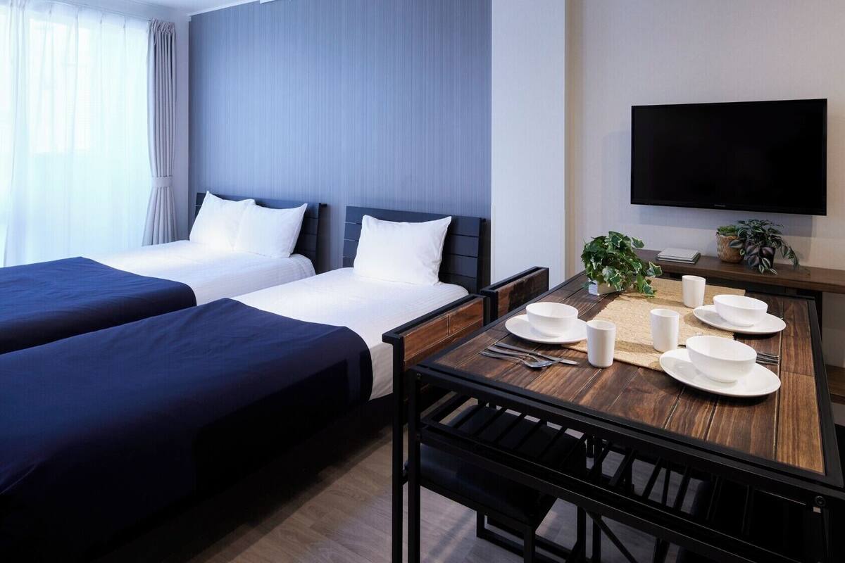 Habitación de hotel con dos camas, una mesa de comedor con platos y cubiertos, un televisor en la pared y una ventana con cortinas.