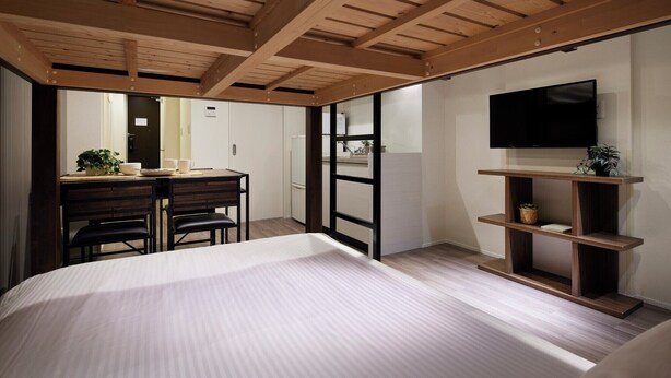 A bedroom with a large bed, a desk with chairs, a television on a shelf, and a sliding door leading to a bathroom. - Minn Kamata (Tokyo)