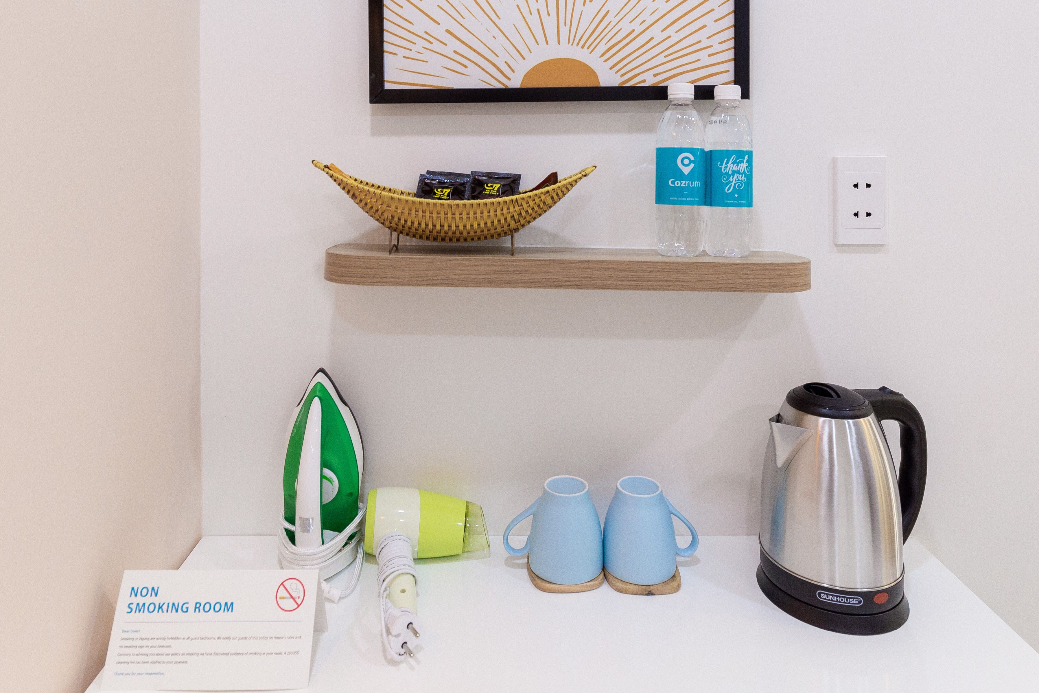 Une chambre d’hôtel avec un panneau « interdiction de fumer », un panier contenant des articles de toilette, deux bouteilles d’eau, une bouilloire et deux tasses sur la table.