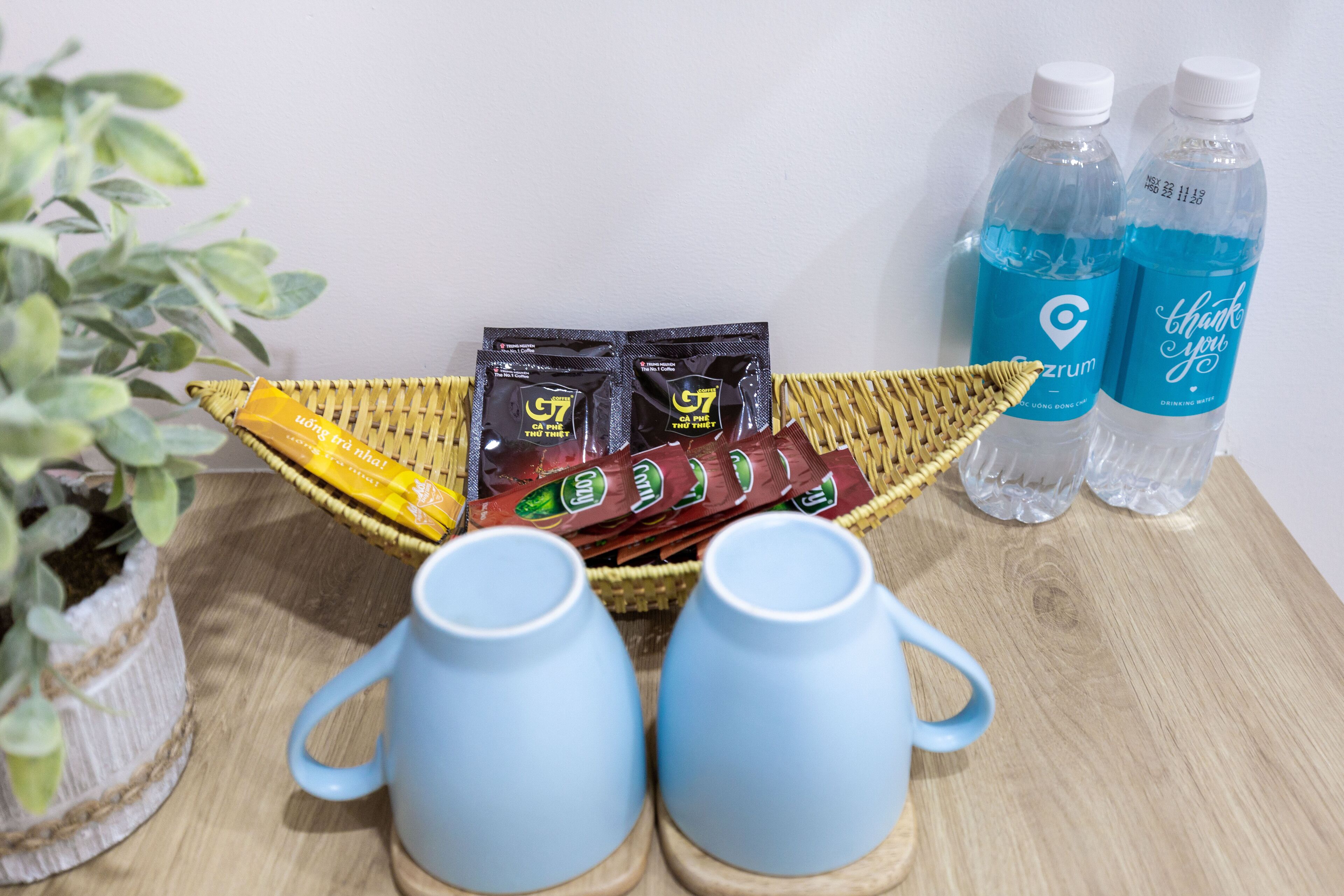 A wicker tray with snack packets, two clear water bottles, and two light blue mugs on a wooden surface.