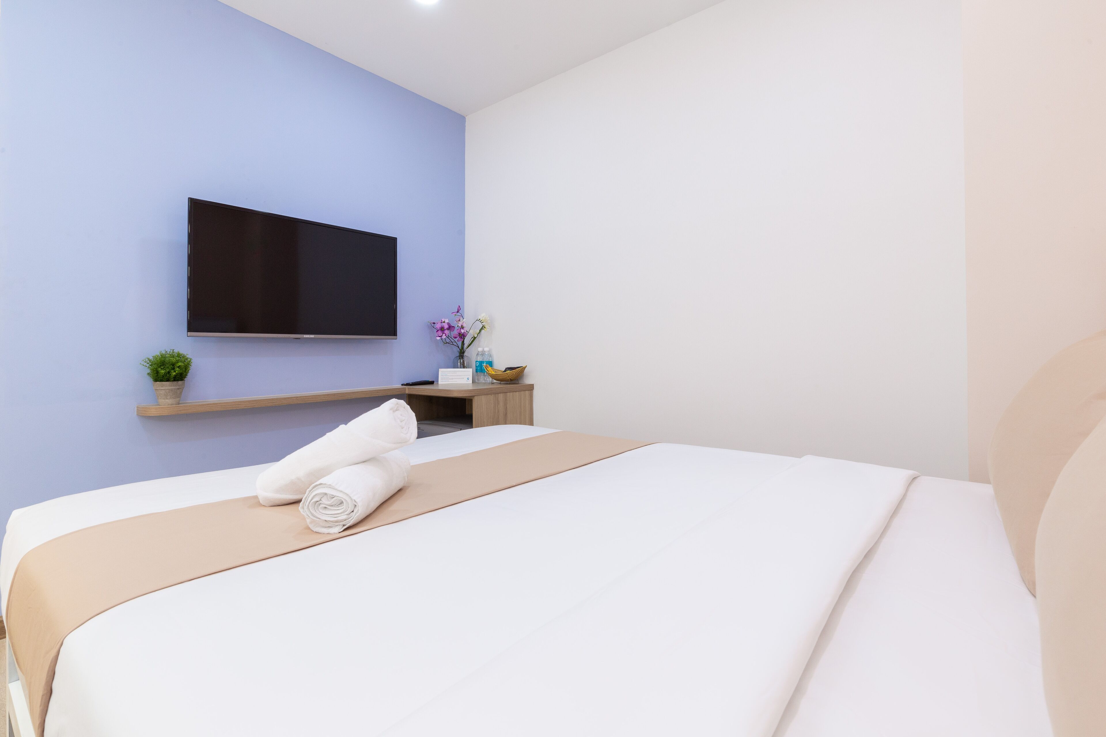 A modern hotel room with a large bed, a flat-screen TV mounted on the wall, a small desk with a plant, and a bedside table with a vase of flowers.