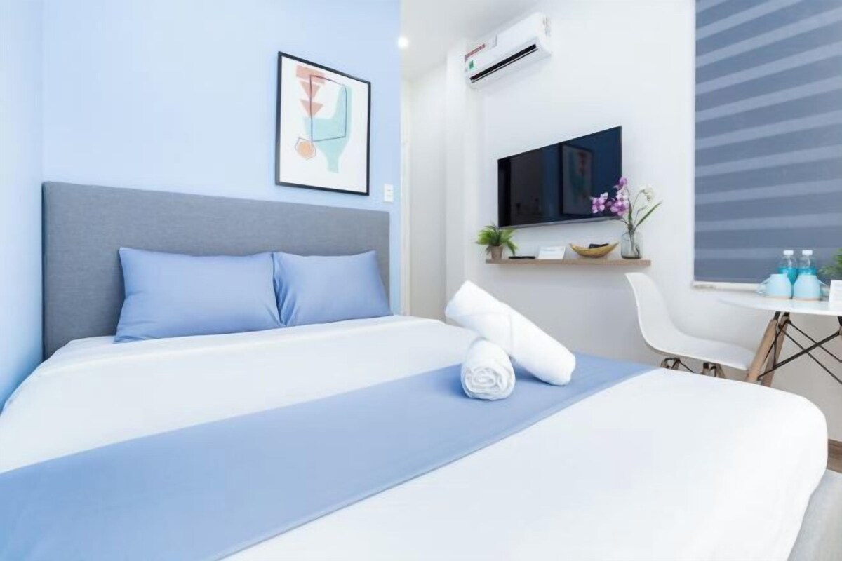 A hotel room with a bed, a TV, a small table with a chair, and a window with blinds.