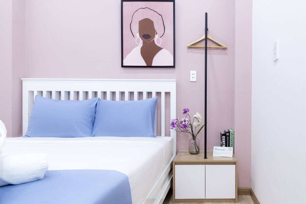 A bedroom with a white bed, blue pillows, a bedside table with a vase of flowers, and a framed picture on the wall.