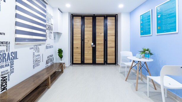 A modern office space with a wooden bench, white chairs, a small table with a plant, and a blue wall with informational posters. - Cozrum Homes Charming Corner (Ho Chi Minh City)