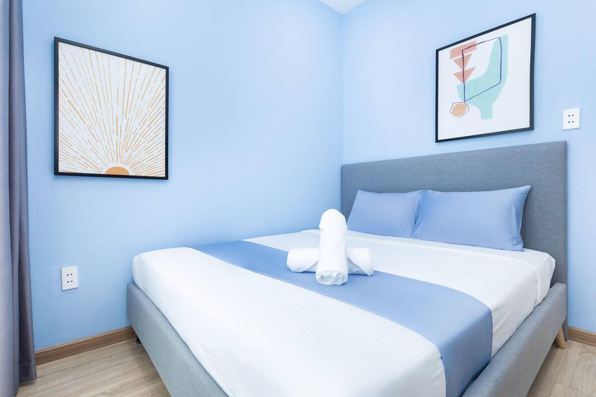 A neatly made bed with a grey headboard, two framed artworks on the walls, and a wooden floor.