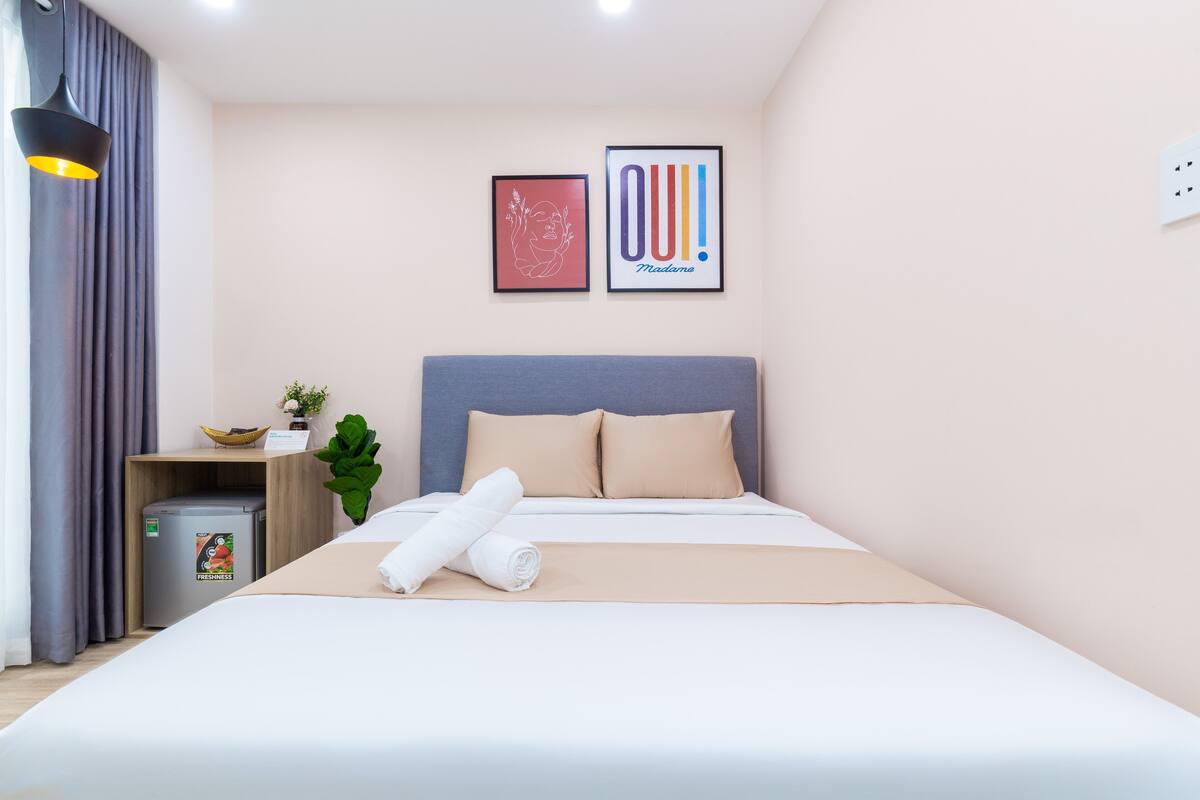 A neatly made bed with a rolled towel, a small side table with a plant, and two framed pictures on the wall.