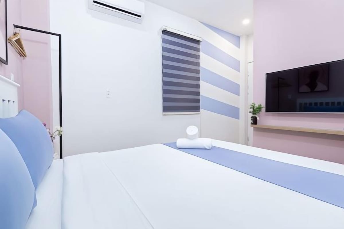A modern bedroom with a bed, a bedside table, a window with blinds, and a wall-mounted air conditioner.