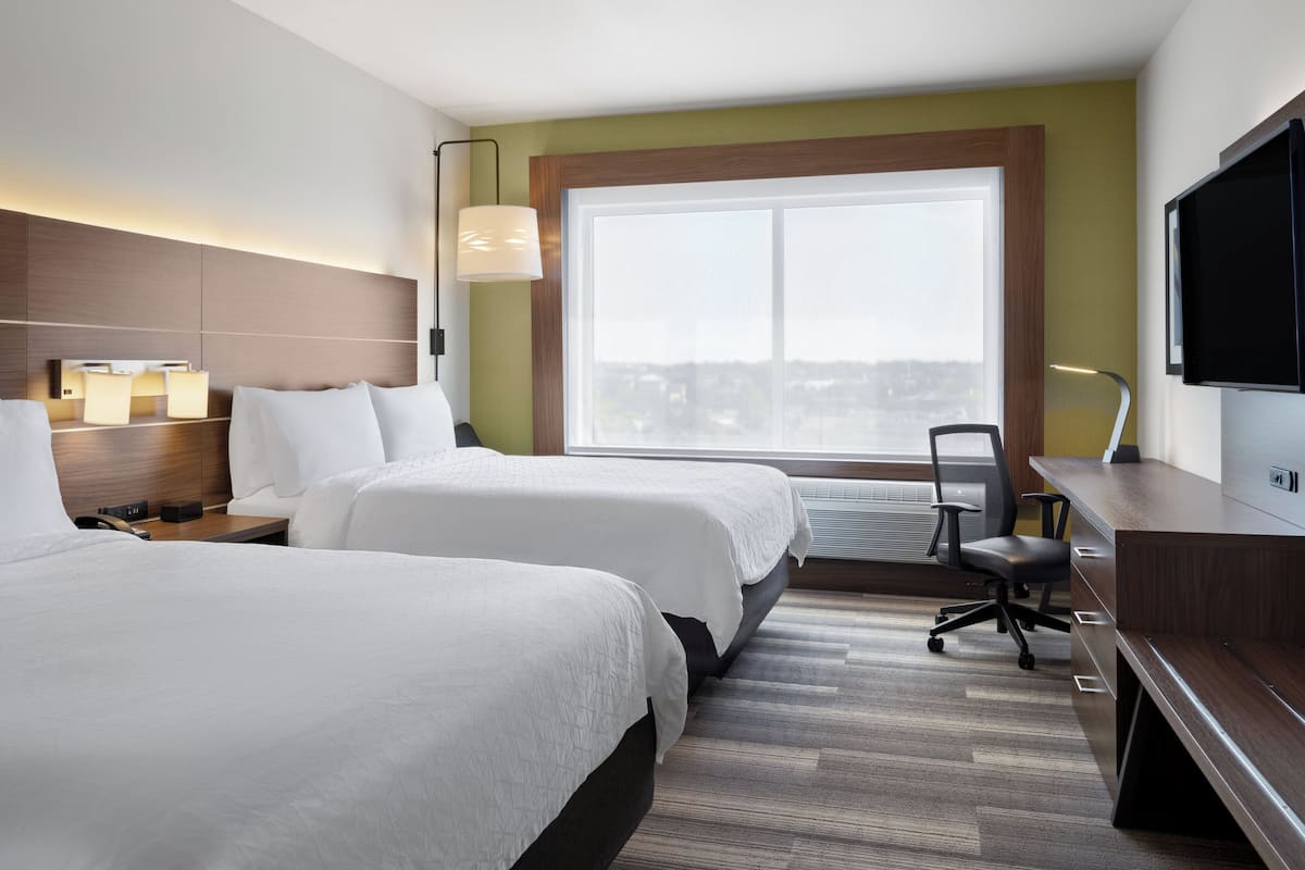 A hotel room with two beds, a desk, a chair, a TV, and a large window with a city view.