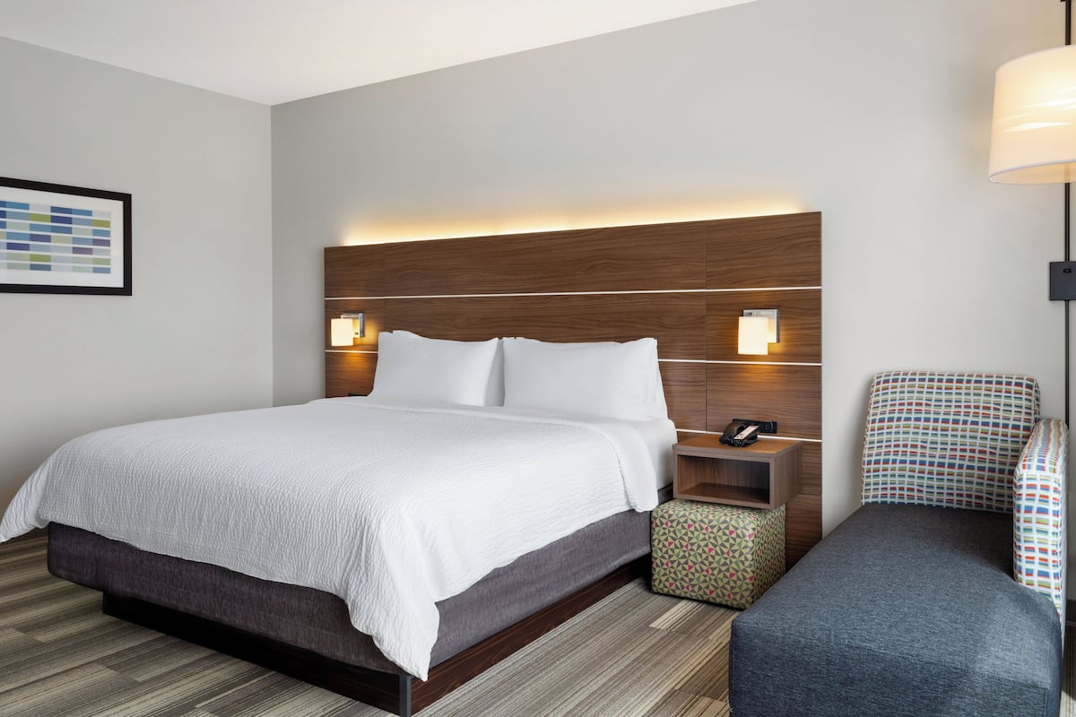 A hotel room with a large bed, bedside tables, a nightstand, a chair, and a small ottoman.