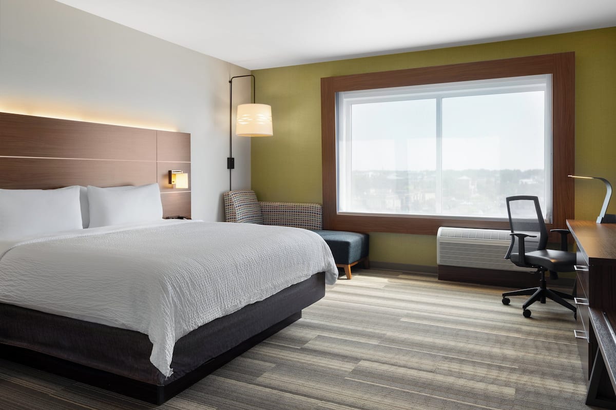 A modern hotel room with a large bed, a desk, a chair, and a large window offering a city view.