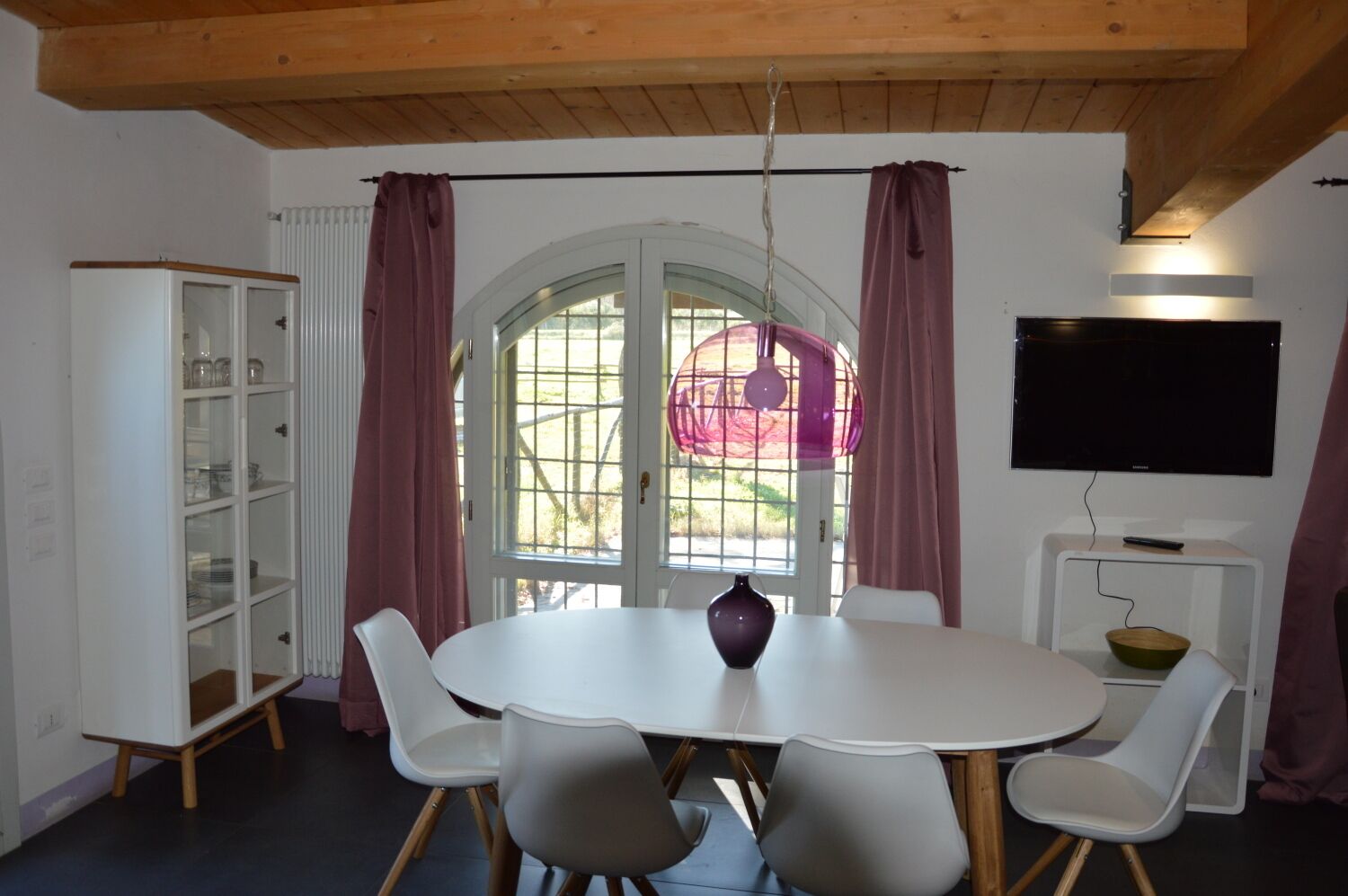 A dining area with a round table, six chairs, a white cabinet with glass doors, a television on a stand, and a window with curtains.