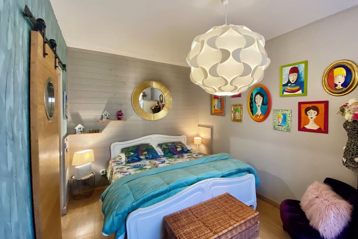 A bedroom with a bed, bedside tables, a hanging light fixture, a mirror, and wall art.