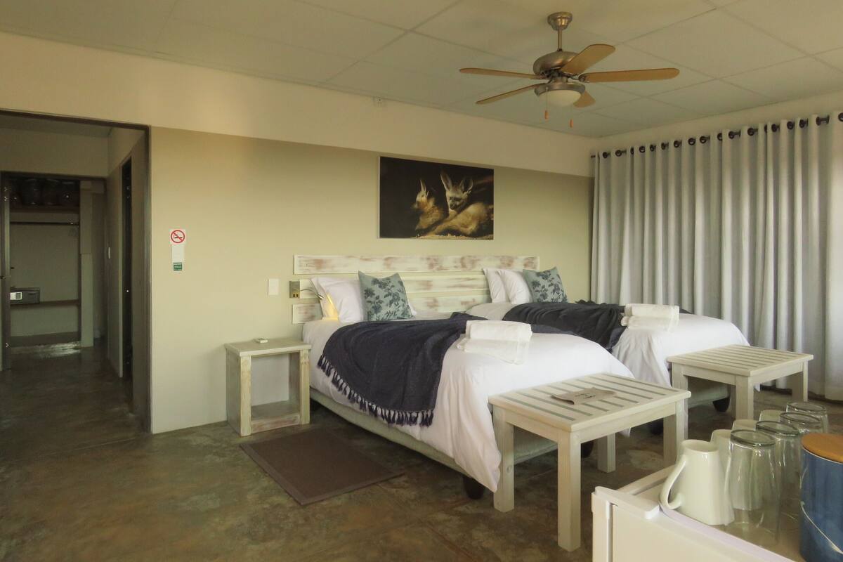 A hotel room with two beds, a ceiling fan, a painting of deer, a nightstand, a bedside table, and a window with curtains.