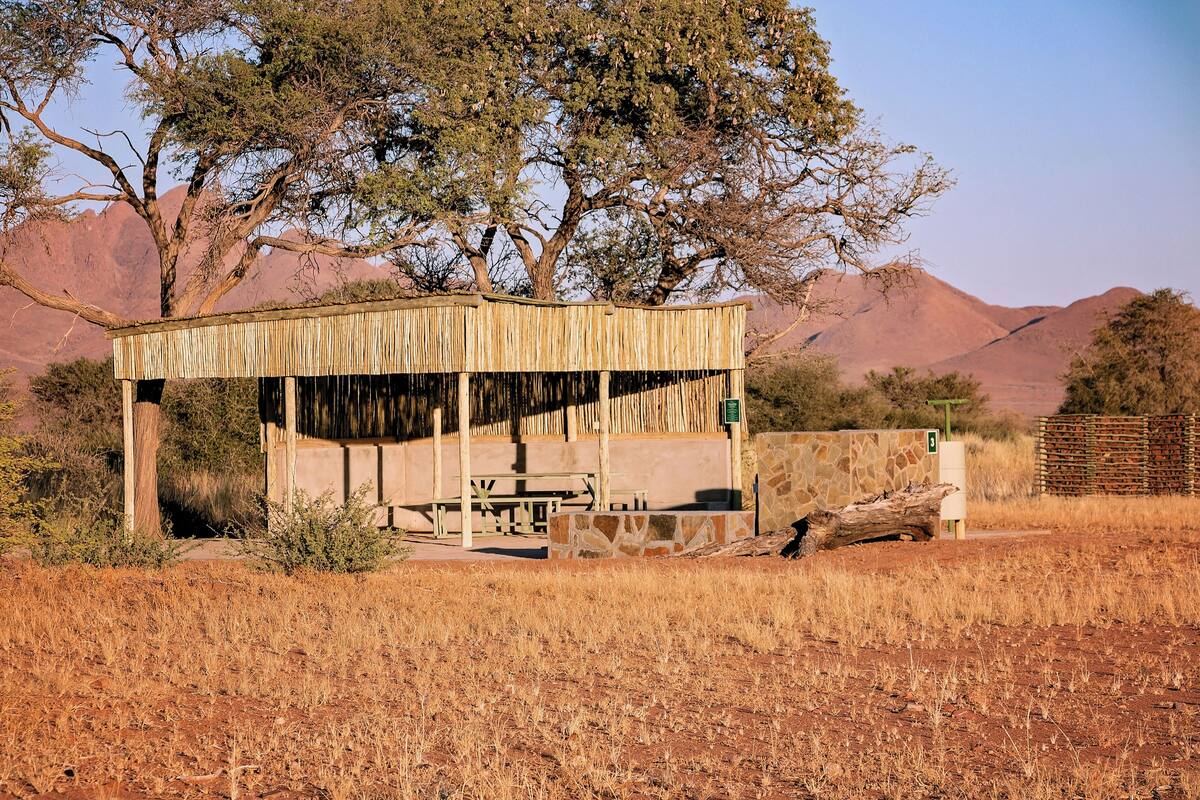 A rustic wooden structure with a bench and picnic table, surrounded by dry grass and trees, set against a mountainous backdrop.