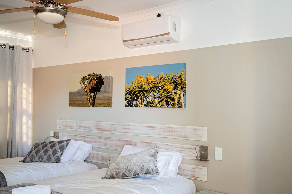 A hotel room with two beds, a ceiling fan, an air conditioning unit, and two paintings on the wall.