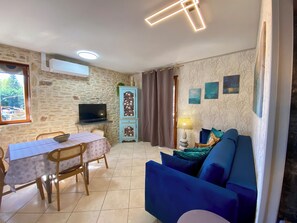 Ein Wohnzimmer mit blauem Sofa, einem Esstisch mit Stühlen, einem Fernseher auf einem Schrank, einem Sideboard und einer Lampe.