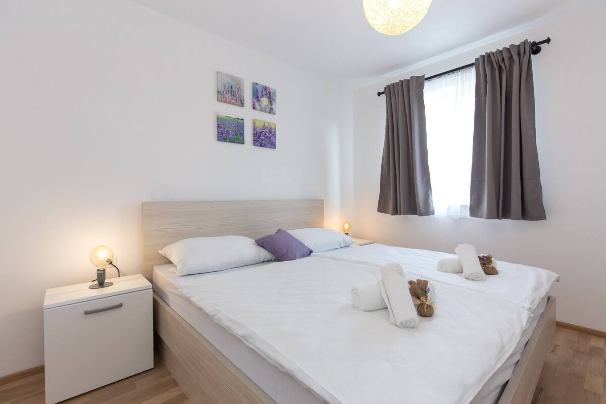 Una cama bien tendida con ropa blanca y un cabecero, una mesita de noche con una lámpara y una ventana con cortinas.