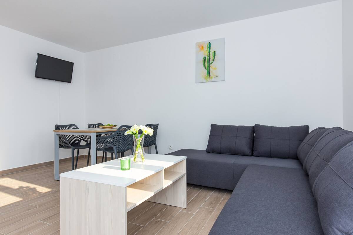 Una sala moderna con un sofá gris, una mesa de centro blanca, un comedor con mesa y sillas, y una pintura de un cactus en la pared.