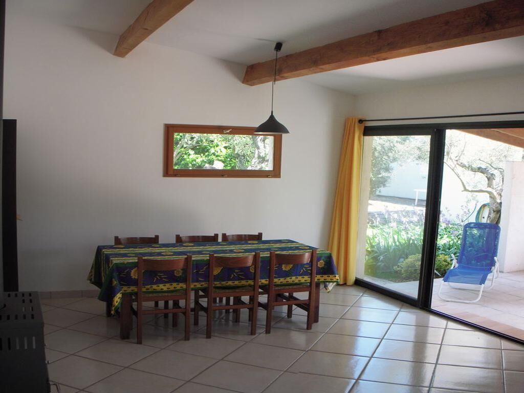 Uma área de refeições com uma mesa posta para seis pessoas, cadeiras de madeira, uma luminária pendente e uma janela com vista para o verde, e uma porta de vidro deslizante que dá para o exterior.