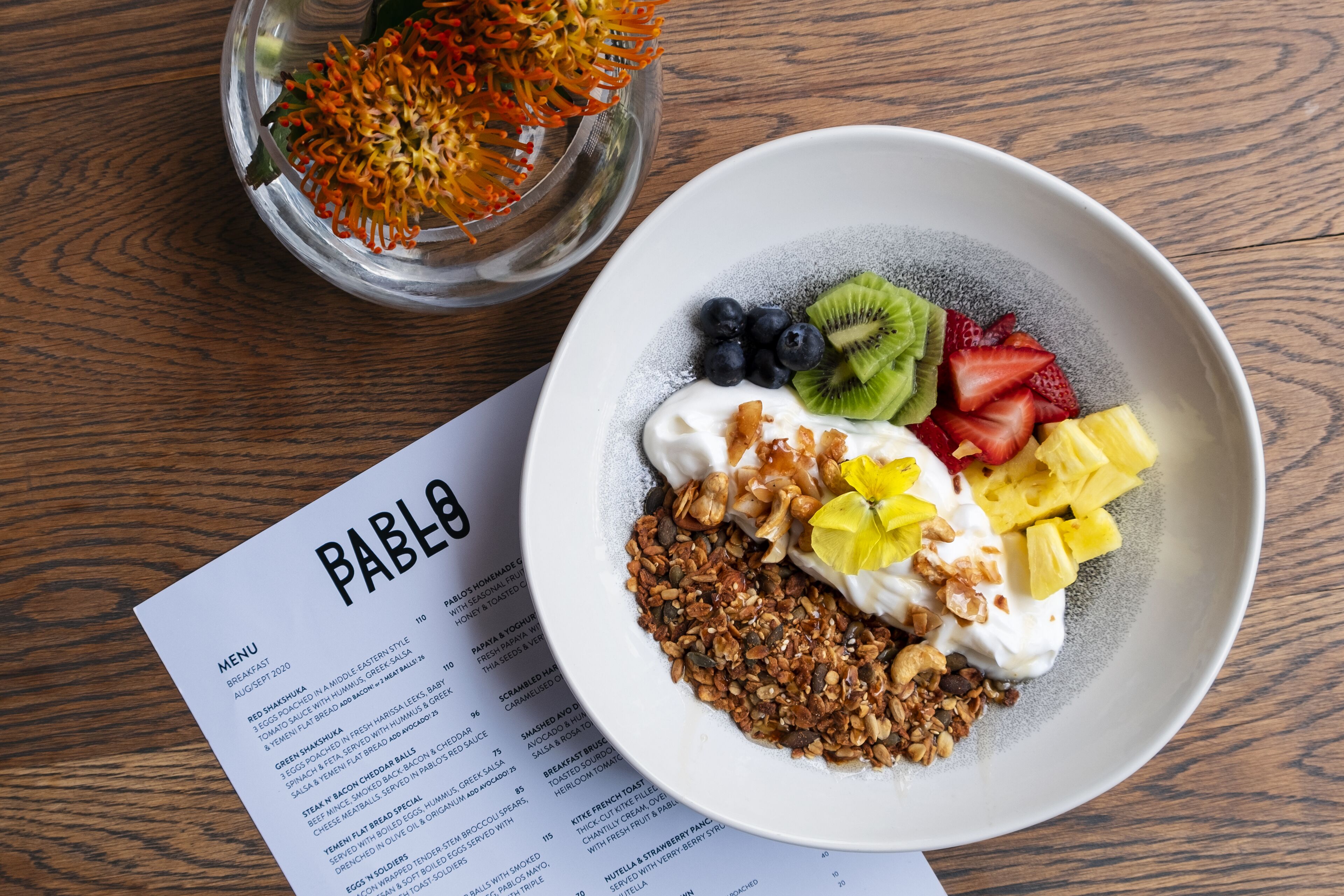 Un bol de yogur con frutas y granola sobre una mesa de madera, con un menú a un lado.
