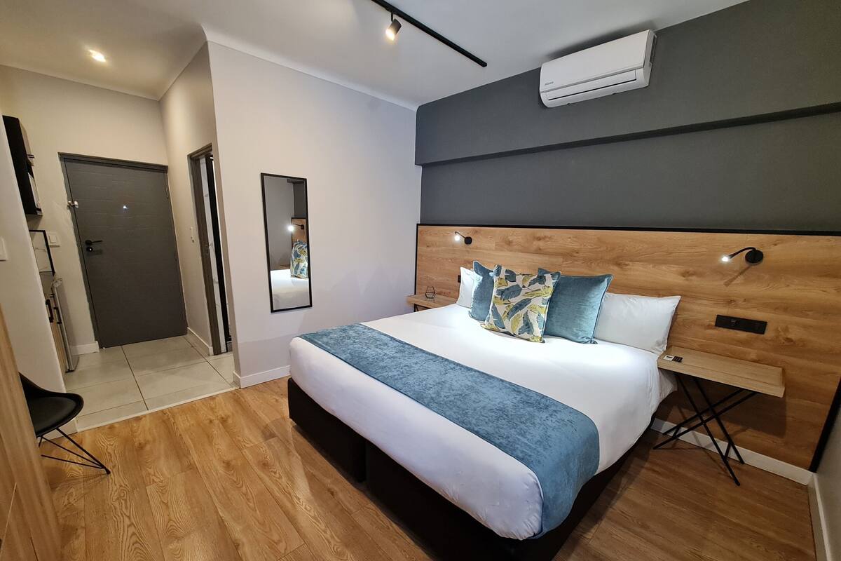 A modern bedroom with a wooden headboard, a large bed with blue and white bedding, a bedside table, and an air conditioning unit on the wall.