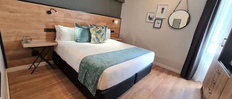 Un dormitorio ordenado con tablero de madera, mesita de noche, cama con manta azul y espejo en la pared.