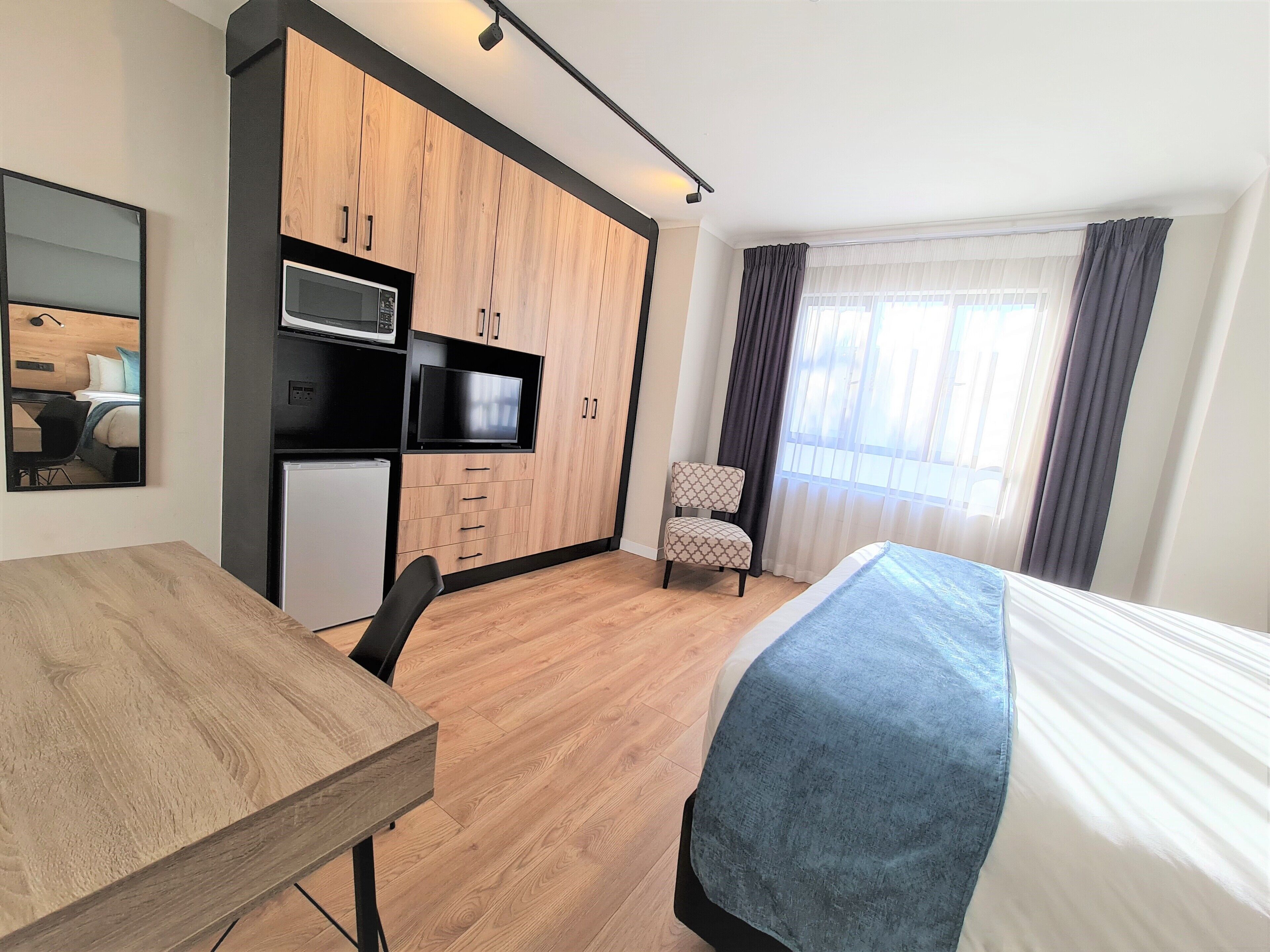 A modern hotel room with a wooden desk, a bed with a blue blanket, a kitchenette with a microwave, and a chair.