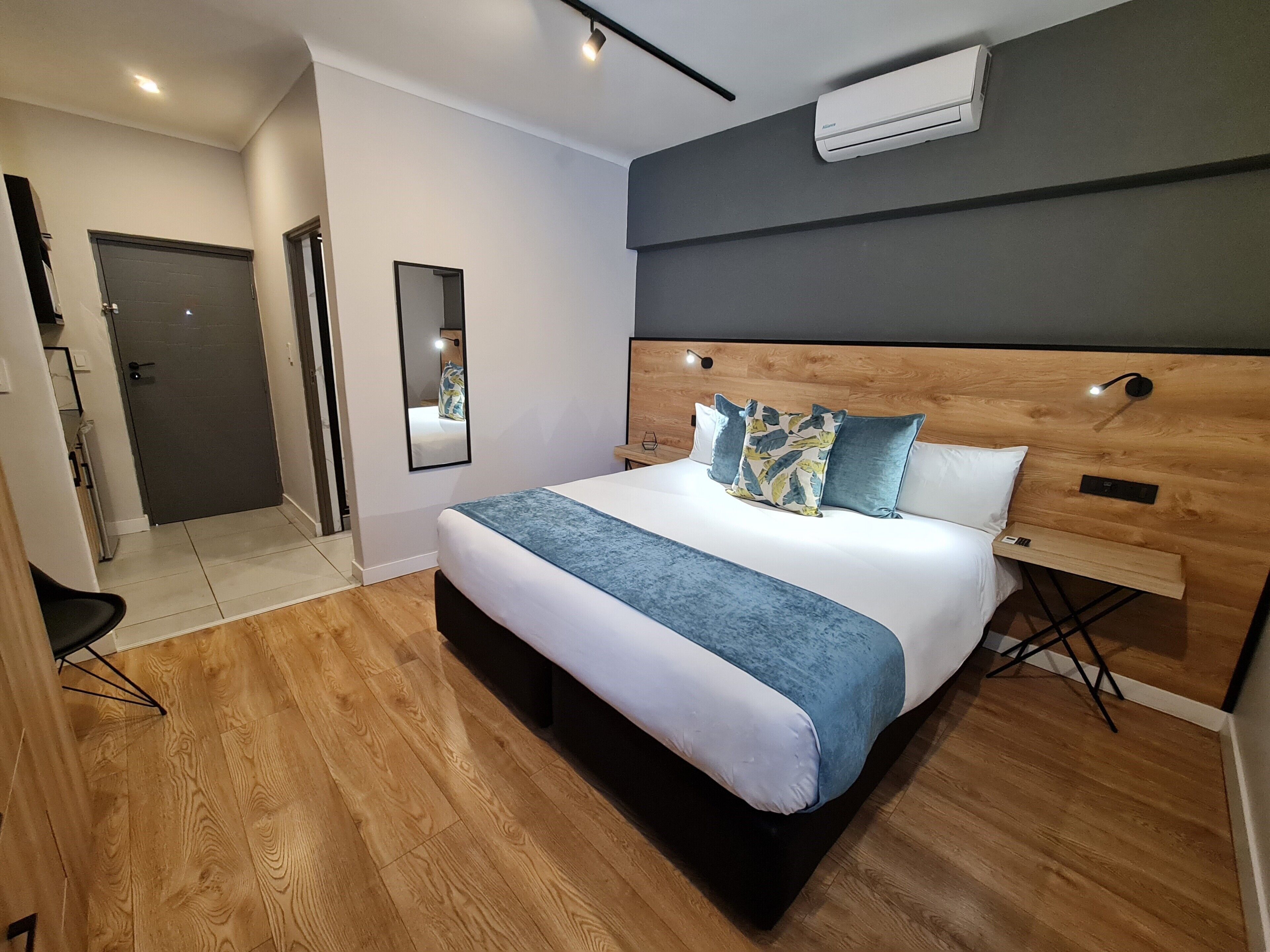 A modern bedroom with a wooden headboard, a large bed with blue and white bedding, a bedside table, and an air conditioning unit on the wall.