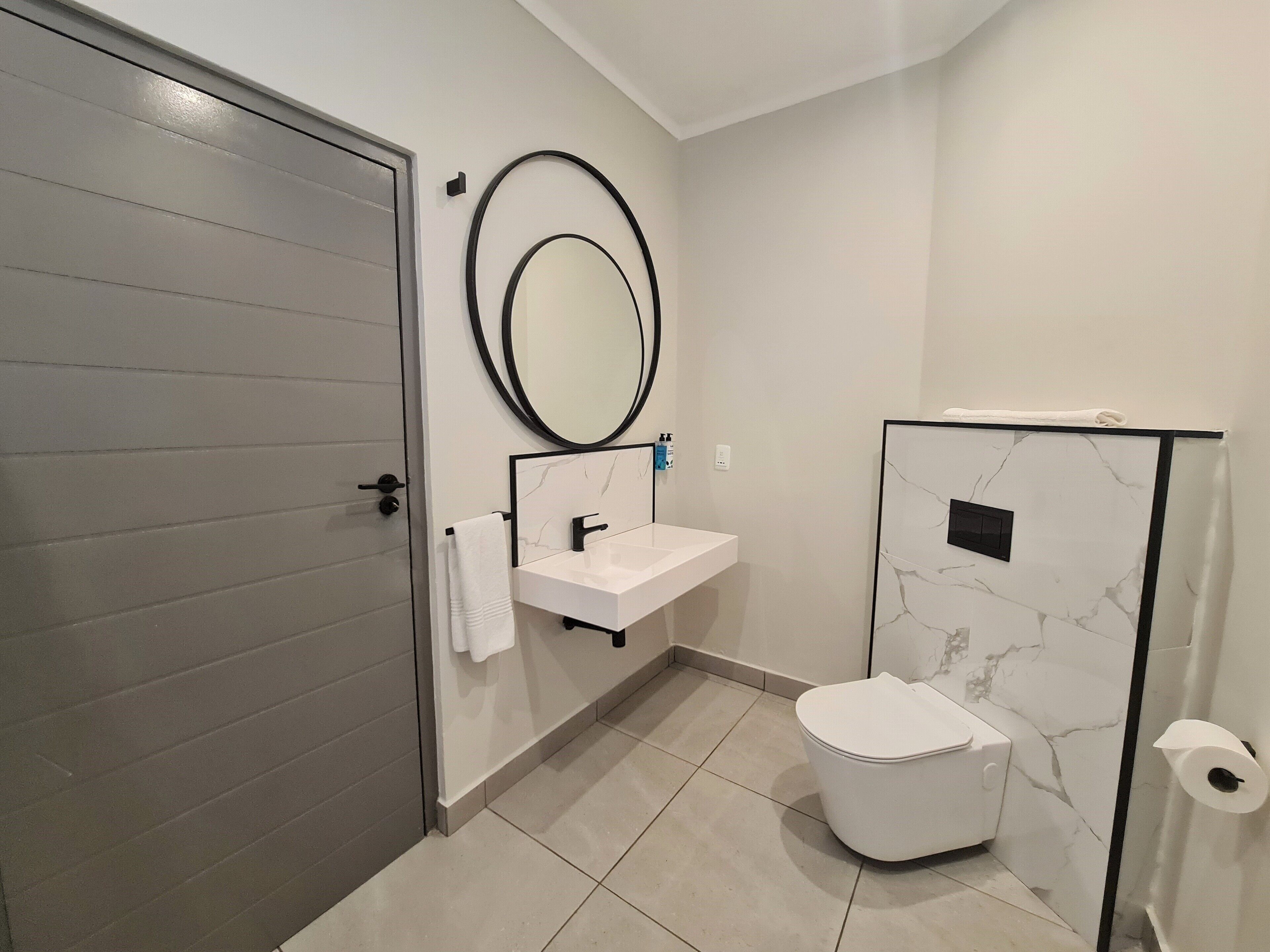 A modern bathroom with a grey door, a white sink with a black faucet, a round mirror, and a toilet with a marble-patterned wall.