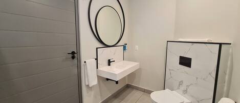 Un baño moderno con una puerta gris, un lavamanos blanco con un grifo negro, un espejo redondo y un inodoro con un revestimiento de pared con diseño de mármol.