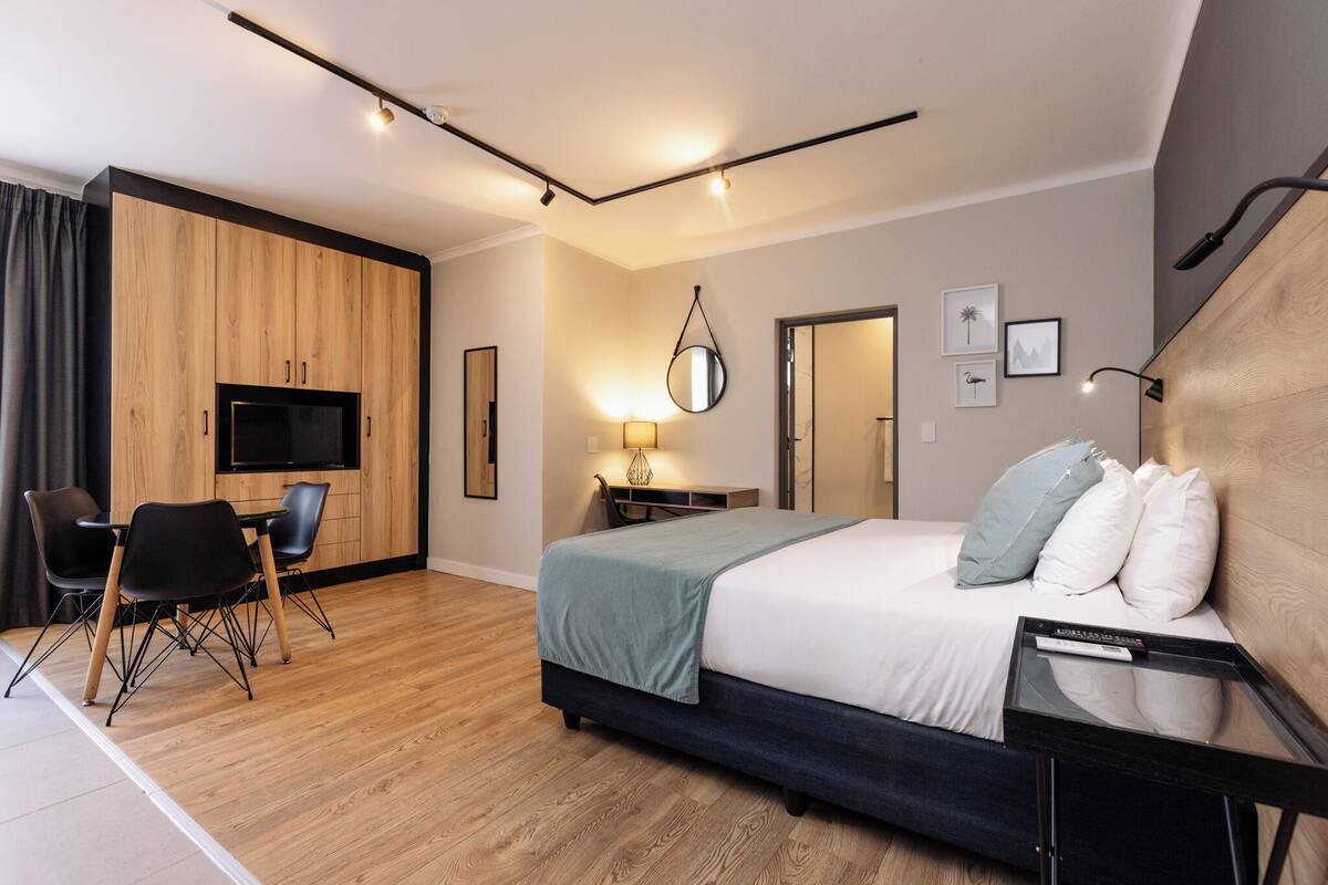 A modern hotel room with a wooden floor, a bed with white and blue pillows, a desk with a lamp, a TV, and a chair.