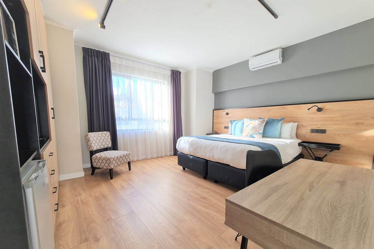 A modern hotel room with a wooden floor, a bed with a wooden headboard, a desk, a chair, and a window with curtains.