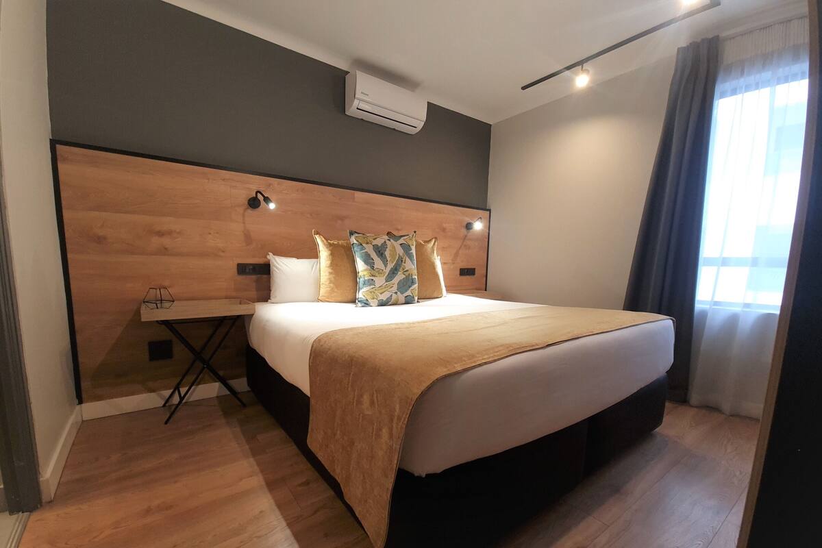 A modern hotel room with a wooden headboard, a bed with a beige blanket, a bedside table with a lamp, and a window with curtains.