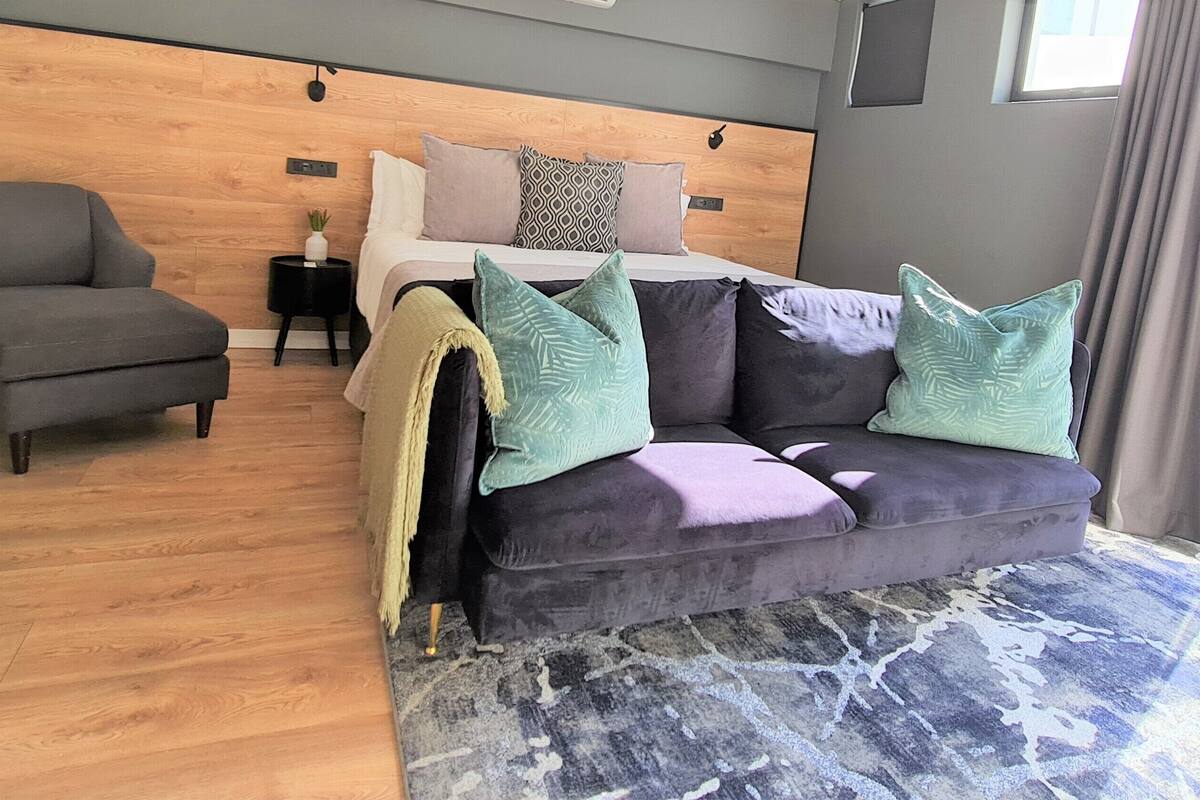 A modern bedroom with a wooden headboard, a grey armchair, a purple sofa with green pillows, and a patterned rug.