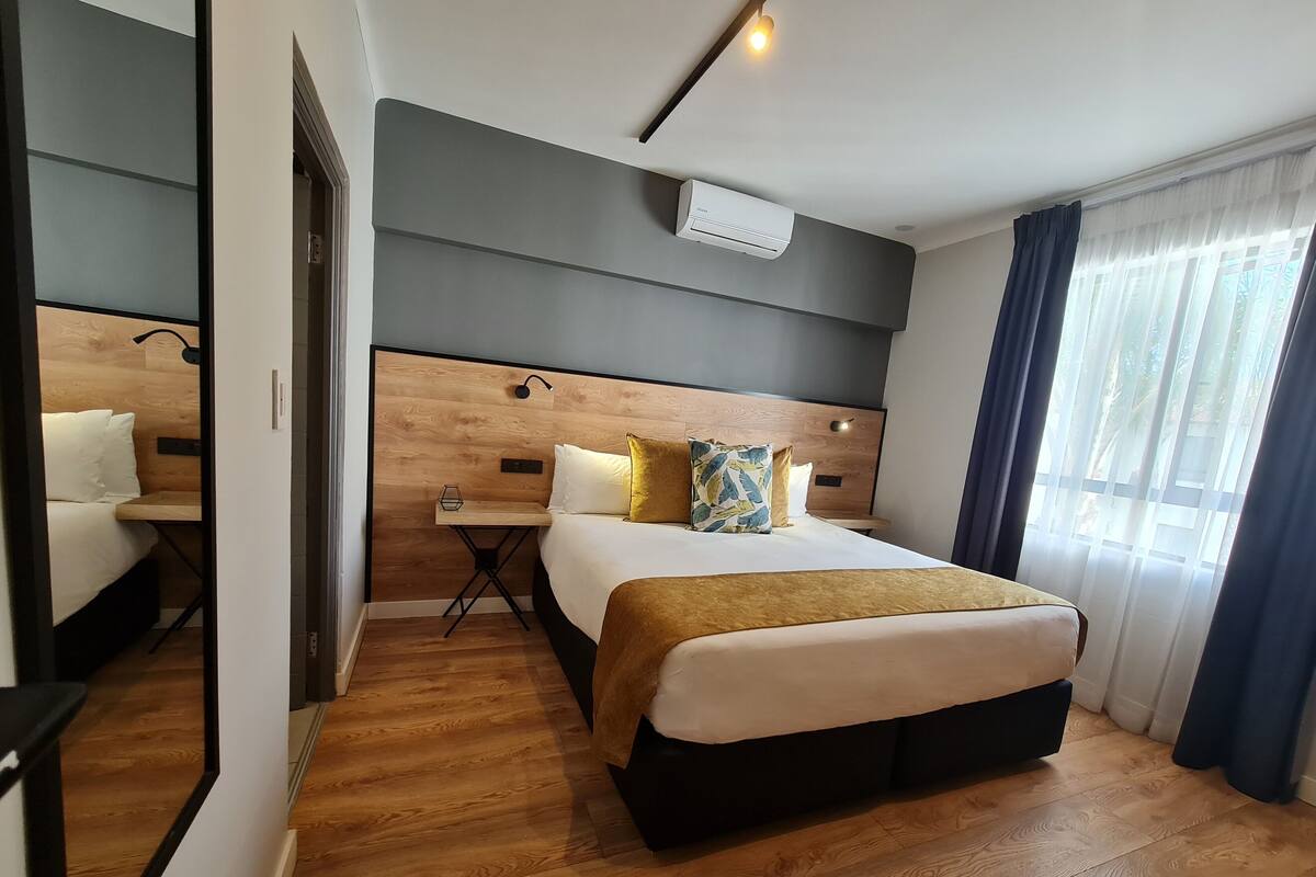 A hotel room with a wooden headboard, a bedside table with a lamp, a bed with a gold throw blanket, and a large mirror.