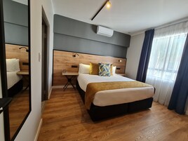 Habitación de hotel con cabecera de madera, una mesita de noche con lámpara, cama con manta dorada y un espejo grande.
