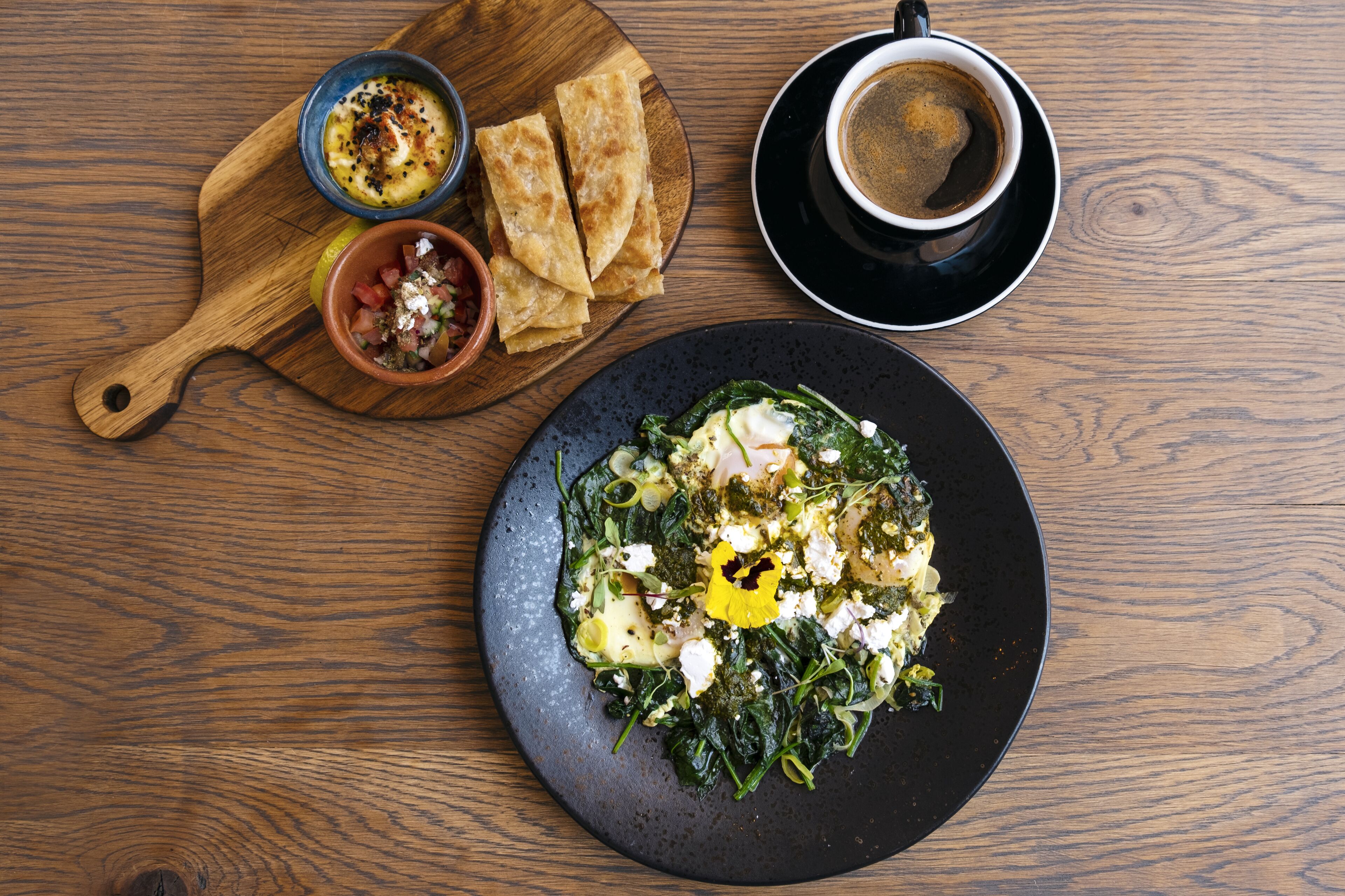 A wooden table with a plate of greens, feta cheese, and a yellow flower garnish, a bowl of tomato and feta, and a cup of coffee on a saucer.