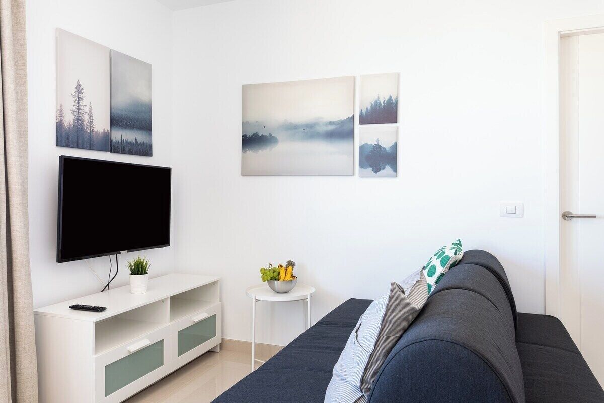 A modern living room with a flat-screen TV on a white stand, a dark grey sofa, and a small white side table with a bowl of fruit.